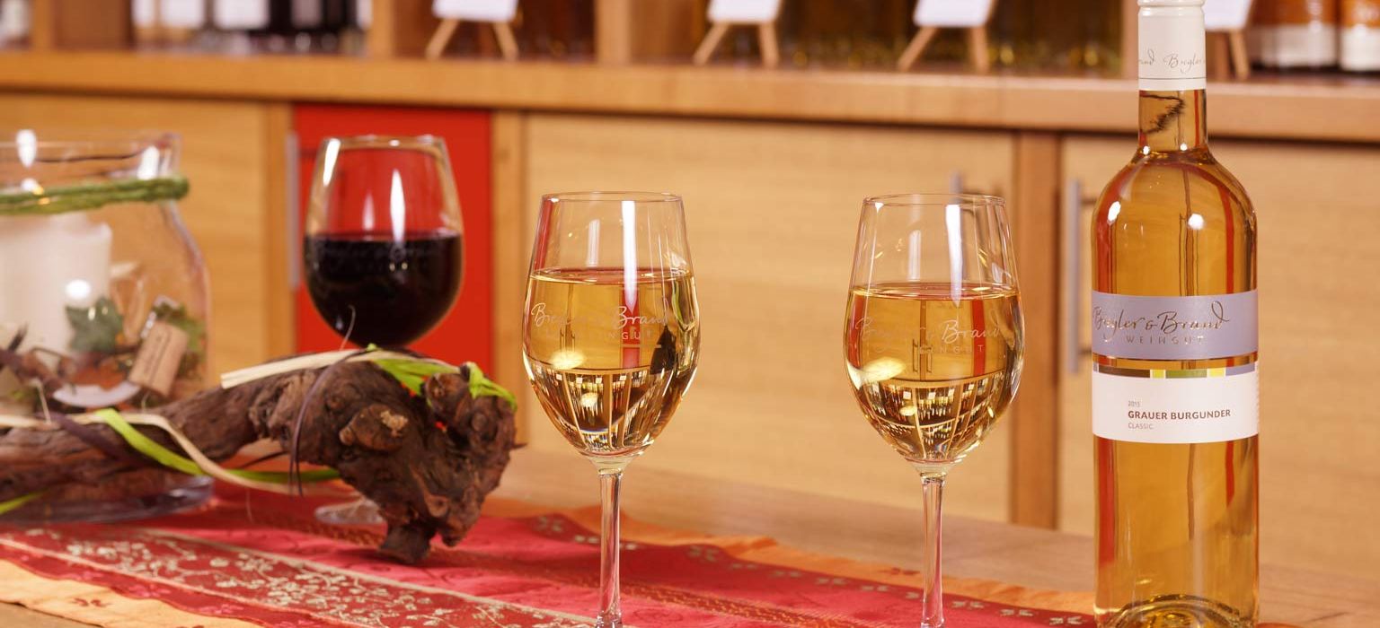 Weinprobe im Weinrestaurant & Vinothek Kaisergarten auf einem Holztisch mit Tischset stehen zwei gefuellte Glas Weisswein, die passende Flasche Wein dazu und ein gefuelltes Glas Rotwein 