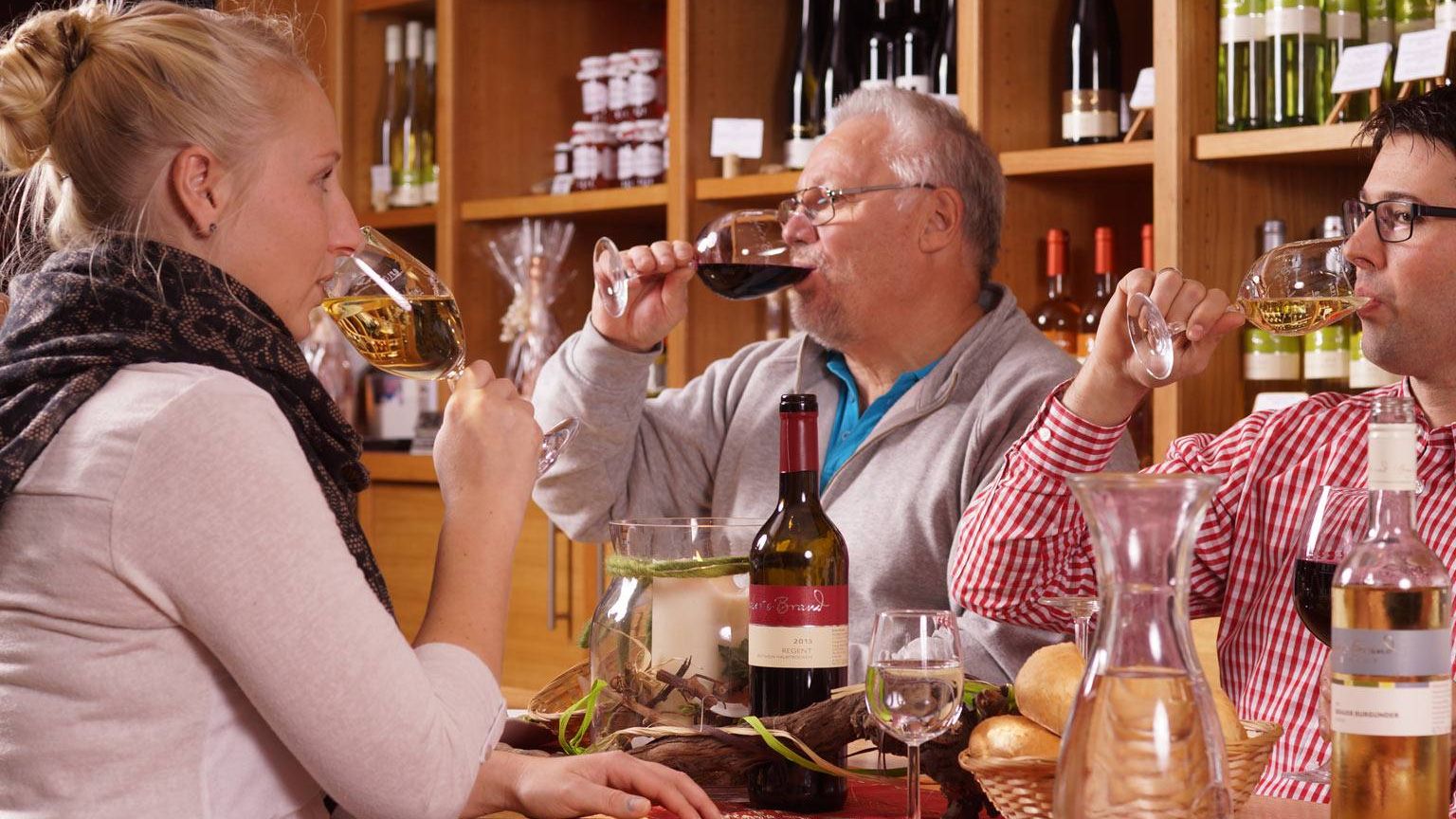 Weinprobe im Weinrestaurant & Vinothek Kaisergarten drei Personen sitzen an einem Holztisch und verkosten die Weine - es stehen Glaeser Flasche Weisswein und Rotwein und eine Karaffe Wasser auf dem Tisch